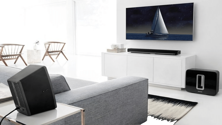 A modern living room with a wall-mounted TV, a soundbar, a subwoofer, and a gray sofa with a projector on the side table.