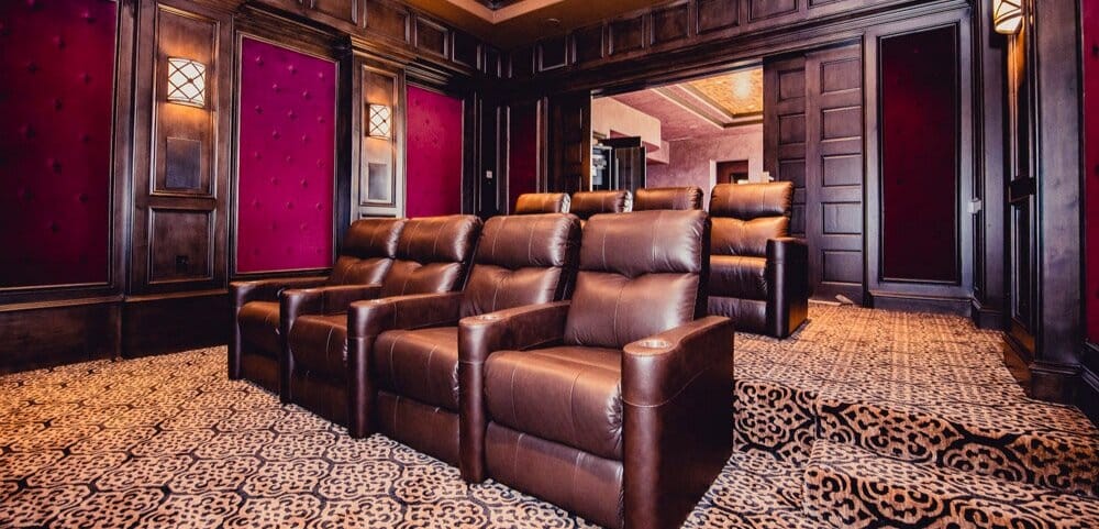 Luxurious home theater room with tiered leather recliners, patterned carpet, and dark wood paneling with red accent walls.