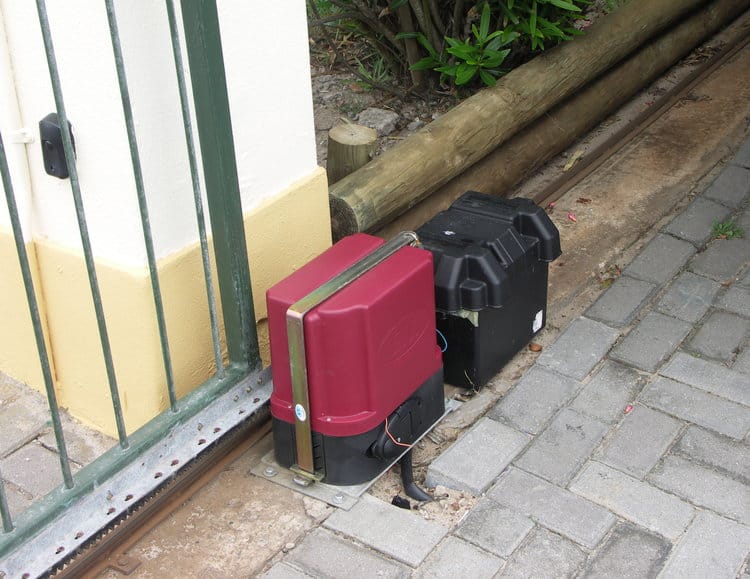 An automated sliding gate motor installed at the base of a metal gate on a paved driveway. The motor housing is red and black, with visible wiring and mounted on a metal plate. Nearby is a black weatherproof enclosure, likely for electrical components. A wooden log fence and greenery are in the background.