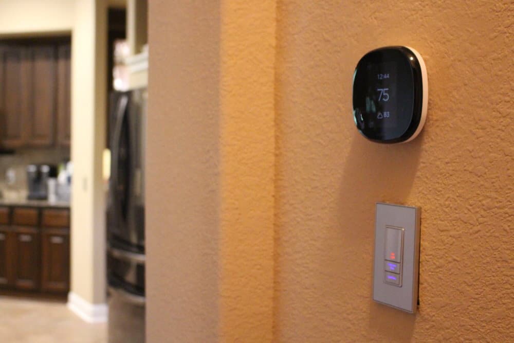 Smart thermostat mounted on a textured wall above an electronic light switch, with a kitchen visible in the background.