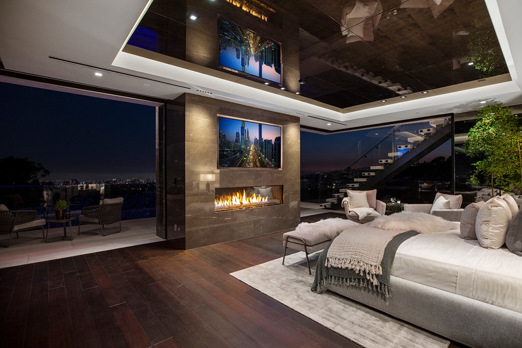 Luxurious modern bedroom with city views at night, featuring a double-sided fireplace, two wall-mounted TVs, a cozy bed, and floor-to-ceiling windows leading to an outdoor balcony and glass staircase.